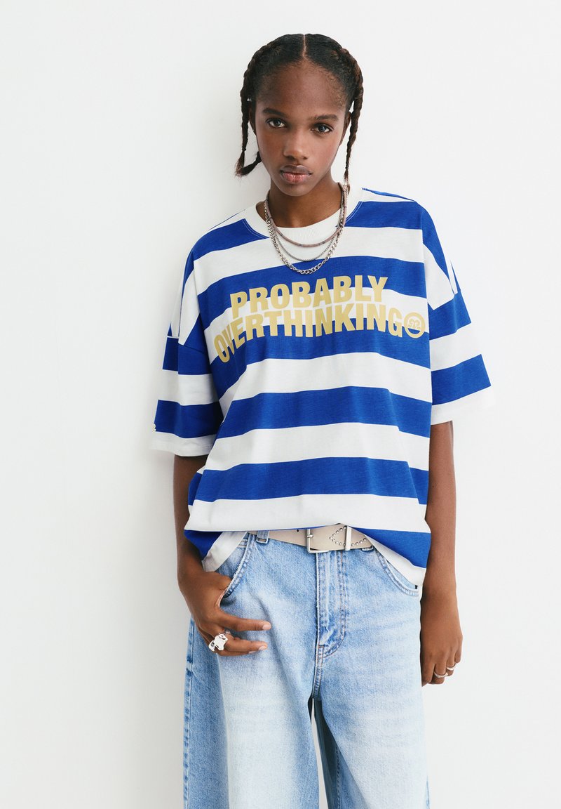 Gestreiftes, übergroßes T-Shirt in Blau und Weiß mit gelbem Text "Wahrscheinlich Überdenken". Kombiniert mit hellblauen Jeansshorts und einem Gürtel.