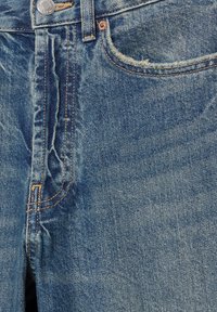 Povečani posnetek modrih denim hlač, ki prikazuje sprednji žep, kovinski gumb in podrobnosti z oranžnim šivom.