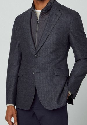 Man in een donkergrijs gestructureerd colbert over een zwarte doorgestikte jas met ritssluiting en marineblauwe broek, met een hand in de zak.