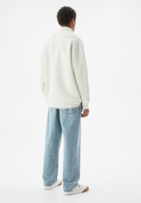 Felpa bianca in fleece con collo alto, abbinata a jeans azzurri larghi e sneakers bianche, vista da dietro.