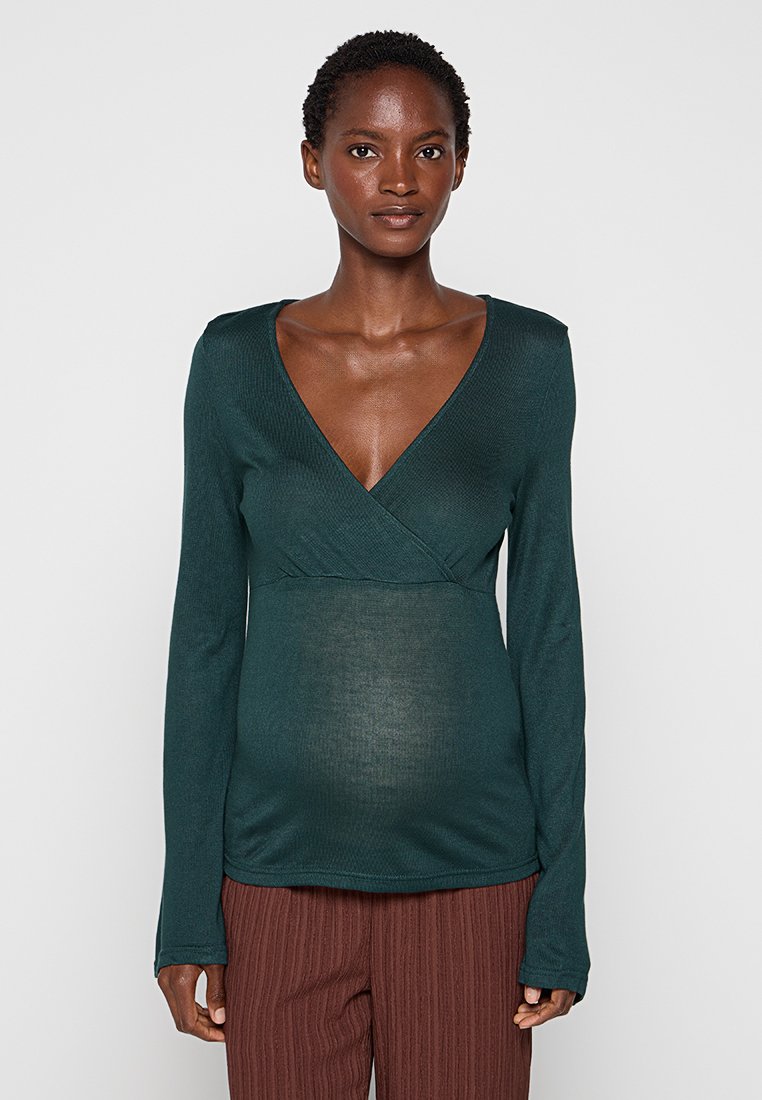 Blusa de manga larga en verde profundo con diseño envolvente y tejido texturizado. Presenta un escote en forma de V y una apariencia suave. Combinada con pantalones marrones a rayas.