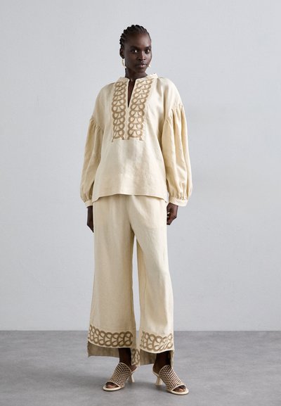 Haut en lin avec une broderie décorative beige et de longues manches bouffantes ; pantalon beige à jambes larges assorti ; sandales à tissage ouvert.