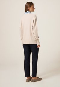 Světle béžový oversized svetr s ribovaným lemem, kombinovaný s tmavě modrými džíny a šedými botami na slip-on. Zadní pohled ukazuje jednoduchý design.