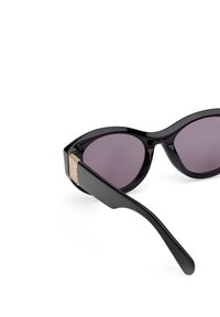 Černé oversized sluneční brýle s fialovými skly, štíhlý plastový rám, odvážný tvar a zlatý akcent na straně.