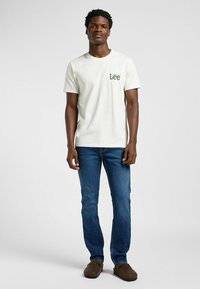 Camisa de algodón blanca con logo verde "Lee", combinada con jeans azules. Modelo usando zapatos de lona marrones; de pie contra un fondo claro.