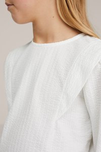 Gros plan d'une personne portant un haut blanc texturé avec des détails larges aux épaules, montrant un profil latéral du cou à la poitrine.