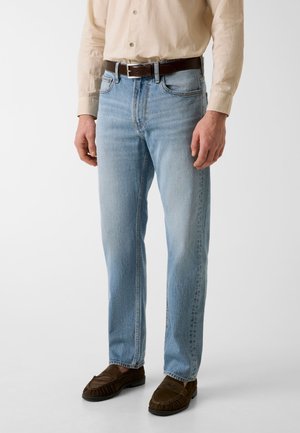 Mann trägt hellblaue Jeans, ein beiges Langarmhemd, eingesteckt, einen dunkelbraunen Gürtel und dunkelbraune Loafers und steht auf einem weißen Boden.