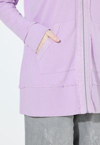 Haut à capuche zippé violet clair avec une bande texturée, poches latérales, ourlet brut et poignets côtelés. Associé à un pantalon gris avec broderies florales.