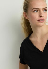 Zwarte V-hals t-shirt met korte mouwen, gemaakt van zachte stof. Model heeft golvend haar en gouden oorringen, tegen een neutrale achtergrond.