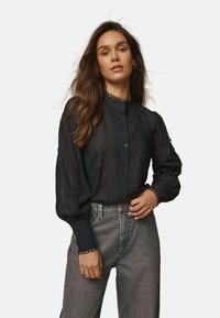 Blusa nera trasparente a maniche lunghe con collo alto, chiusura a bottoni e dettagli in pizzo decorativo sulle maniche e sul collo.