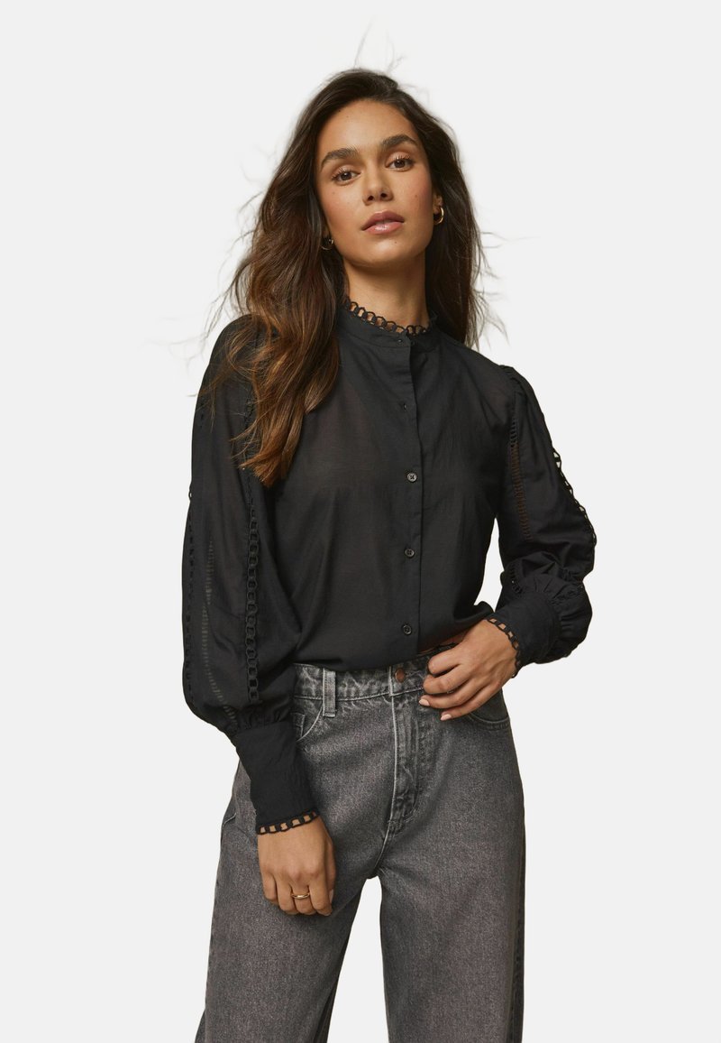 Blusa nera trasparente a maniche lunghe con collo alto, chiusura a bottoni e dettagli in pizzo decorativo sulle maniche e sul collo.