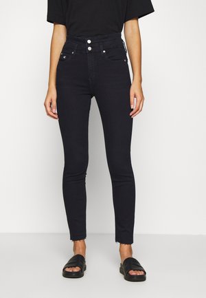 Osoba nosząca czarne, dopasowane dżinsy z wysokim stanem i postrzępionymi nogawkami oraz czarne klapki z odkrytymi palcami, stojąca na białym tle.