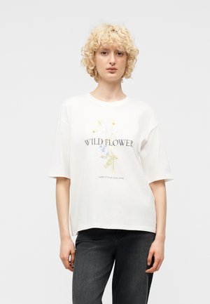 Person med krøllet blondt hår iført en hvid oversized T-shirt med blomsterprint og teksten "WILD FLOWER", kombineret med sorte jeans.