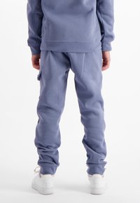 Persoon in blauwe cargopantalons en bijpassende sweatshirt, staand met ontspannen handen, witte Nike Air sneakers zichtbaar van achter.