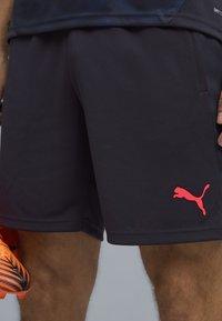 Shorts deportivos negros con tejido texturizado, bolsillos laterales y un destacado logo rosa de Puma en el dobladillo izquierdo.