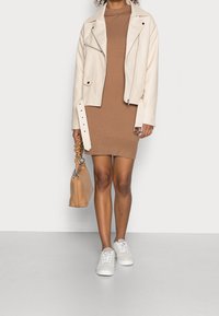 Veste en cuir crémeuse de style motard portée sur une robe marron cintrée, associée à des baskets blanches. Comprend un sac à main beige avec une anse froncée.