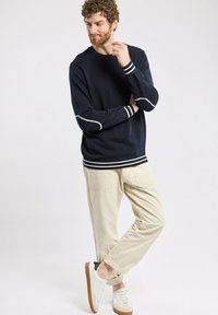 Sweat-shirt marine avec des poignets et un ourlet à rayures blanches, associé à un pantalon beige et des baskets blanches. Tissu lisse, coupe d�écontractée, détails minimaux.