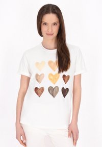 Hvid T-shirt med trykt design af ni hjerter i forskellige nuancer af beige og brun, præsenteret i et gittermønster. Korte ærmer.