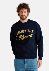 Marineblauwe sweatshirt met goud geborduurde tekst "ENJOY THE MOMENT", ronde hals, lange mouwen en een relaxte pasvorm, gecombineerd met lichtblauwe jeans.