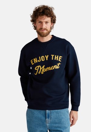 Marineblauwe sweatshirt met goud geborduurde tekst "ENJOY THE MOMENT", ronde hals, lange mouwen en een relaxte pasvorm, gecombineerd met lichtblauwe jeans.