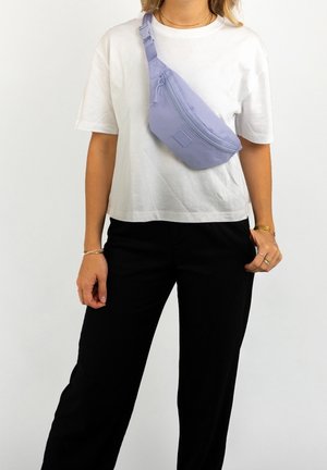 Vrouw met een wit T-shirt en zwarte broek, met een lichtpaarse schoudertas over de schouder, tegen een witte achtergrond.