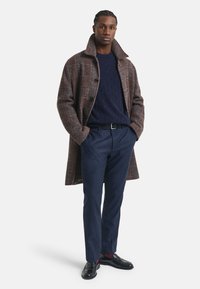Brauner kariert Wollmantel, navyblauer Strickpullover, dunkle Hose, schwarze Schuhe und Gürtel. Insgesamt strukturierte Silhouette und kontrastierende Texturen.