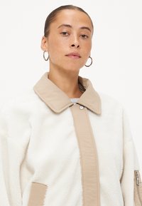 Neplederen jas in crèmekleur met een beige, overgrote kraag en ritssluitingaccent. Zachte textuur en vrijblijvende pasvorm met drukknopen sluiting.