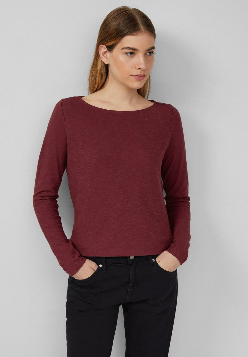 Bordeaux langärmliges T-Shirt mit Rundhalsausschnitt und strukturiertem Stoff, kombiniert mit schwarzen Jeans, die Hände in den Taschen. Einfaches Design.