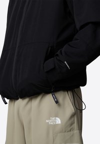 Personne portant une veste noire zippée avec le texte "WindWall" sur la manche et un pantalon beige avec le logo "The North Face" sur la cuisse.