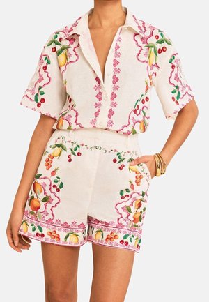 Kort hvid skjorte med knapper og matchende shorts med farverige frugt- og pink blomsterprint, båret med guldbånd på det ene håndled.