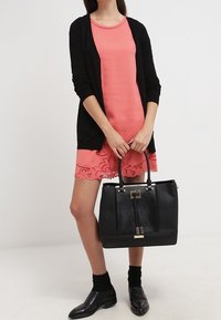 Černá kabelka se strukturovaným designem, zlatými kovovými detaily a texturovaným povrchem. Model nosí korálové šaty s krajkovými detaily, černý cardigan a boty.