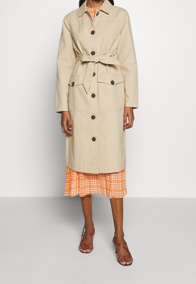 Femme portant un trench-coat beige ceinturé par-dessus une robe plissée à carreaux orange, associée à des sandales à talons marron avec des brides.