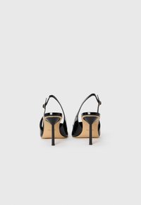 Un paio di scarpe con tacco alto in vernice nera, modello slingback, con tacchi a stiletto sottili, viste da dietro su una superficie bianca.