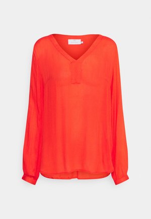 Blouse rouge vif à manches longues avec encolure en V et manches retroussées, coupe ample, présentée sur un fond gris clair.