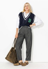 Gilet en tricot marine avec un patch vert "B" sur une chemise blanche, associé à un pantalon taille haute à carreaux gris et à des chaussures à enfiler à imprimé léopard. Sac marron.