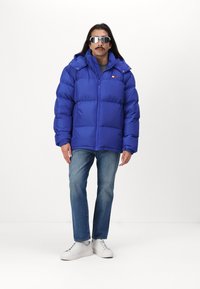 Chaqueta acolchada azul con capucha, con cierre de cremallera y un pequeño logo. Combinada con jeans de un azul claro y zapatillas blancas. Textura suave.