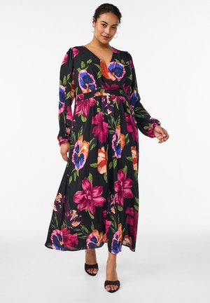 Kvinde iført en sort maxikjole med lange ærmer og store farverige blomsterprint, samt sorte åbne hæle, stående foran en hvid baggrund.