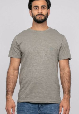 Homme aux cheveux courts et foncés avec une barbe, portant un t-shirt gris uni à manches courtes et un jean bleu, debout devant un fond uni.