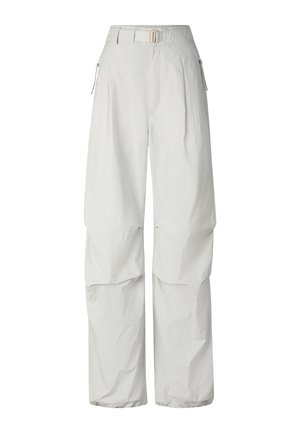 Femme aux cheveux blonds et lunettes de soleil, portant un haut court noir noué, un pantalon blanc ample et des baskets blanches, debout devant un fond blanc.