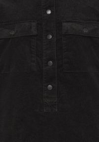 Cămașă neagră din catifea cu două buzunare la piept, nasturi metalici negri pe fanta centrală și o textură a țesăturii.