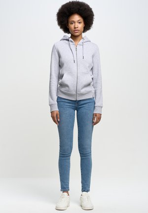 Sudadera gris con cremallera y bolsillo tipo canguro, capucha con cordón y puños acanalados. Combinada con unos vaqueros ajustados azules y zapatillas blancas.