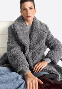 Sivý medvedí kabát s textúrovanou tkaninou, veľkými klopami a modrými manžetovými rukávmi. Modelka má na sebe sivý sveter a sedí s hnedou taškou.