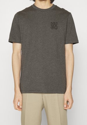 Person iført en mørkegrå kortærmet T-shirt med "HUGO"-logoet på øverste venstre del af brystet og lyse beige bukser.