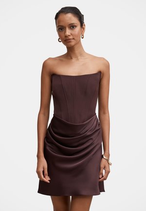 Femme portant une robe bustier marron foncé avec des détails drapés sur la jupe, debout les mains relâchées le long du corps.