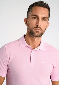 Polo de color rosa claro en punto texturizado, con cuello blanco y tapeta de botones de dos, y un pequeño logotipo blanco en el pecho.