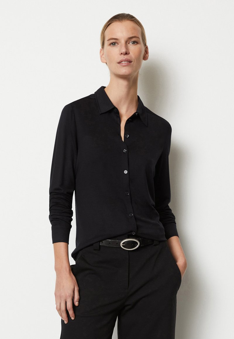 Camisa de botón de manga larga negra con cuello, hecha de material suave. Presenta una textura sutil y un cinturón negro con una hebilla circular plateada.