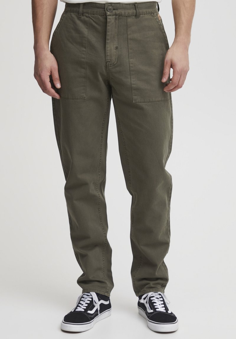 Pantalon cargo vert en coton avec deux poches avant, une coupe avant plate et une coupe droite au niveau des jambes. Porté avec des baskets noires avec des accents blancs.