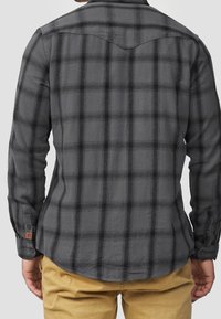 Chemise en flanelle grise avec motif à carreaux noirs, manches longues et poignets à boutons. Présente un ourlet légèrement courbé et un empiècement au dos.