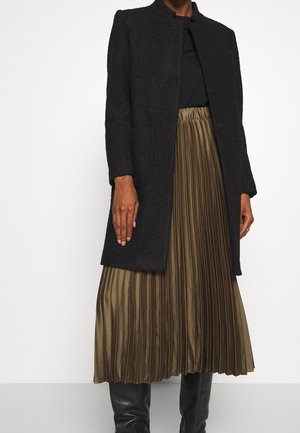 Vrouw draagt een zwarte textuurlagen jas over een zwart topje en een olijfgroene geplooide midi-rok met zwarte laarzen tot net onder de knie.