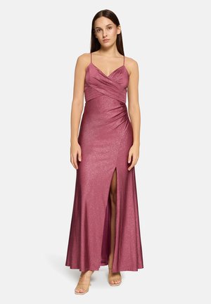 Kvinde står, iført en ærmeløs, glitrende lyserød aftenkjole med tynde stropper og en front-skråskæring helt op til låret, kombineret med sandaler med klare hæle.